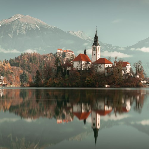 A rustic village rest on the edge of a mirror lake, a massive mountain looms behind.