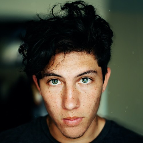Portrait of a man with tossled black hair and prominent freckles gazing meaningfully into the camera.